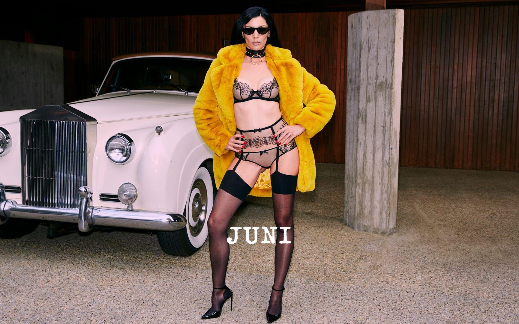 A brunette model stands with hands on hips in front of a car, wearing the black Juni lingerie set